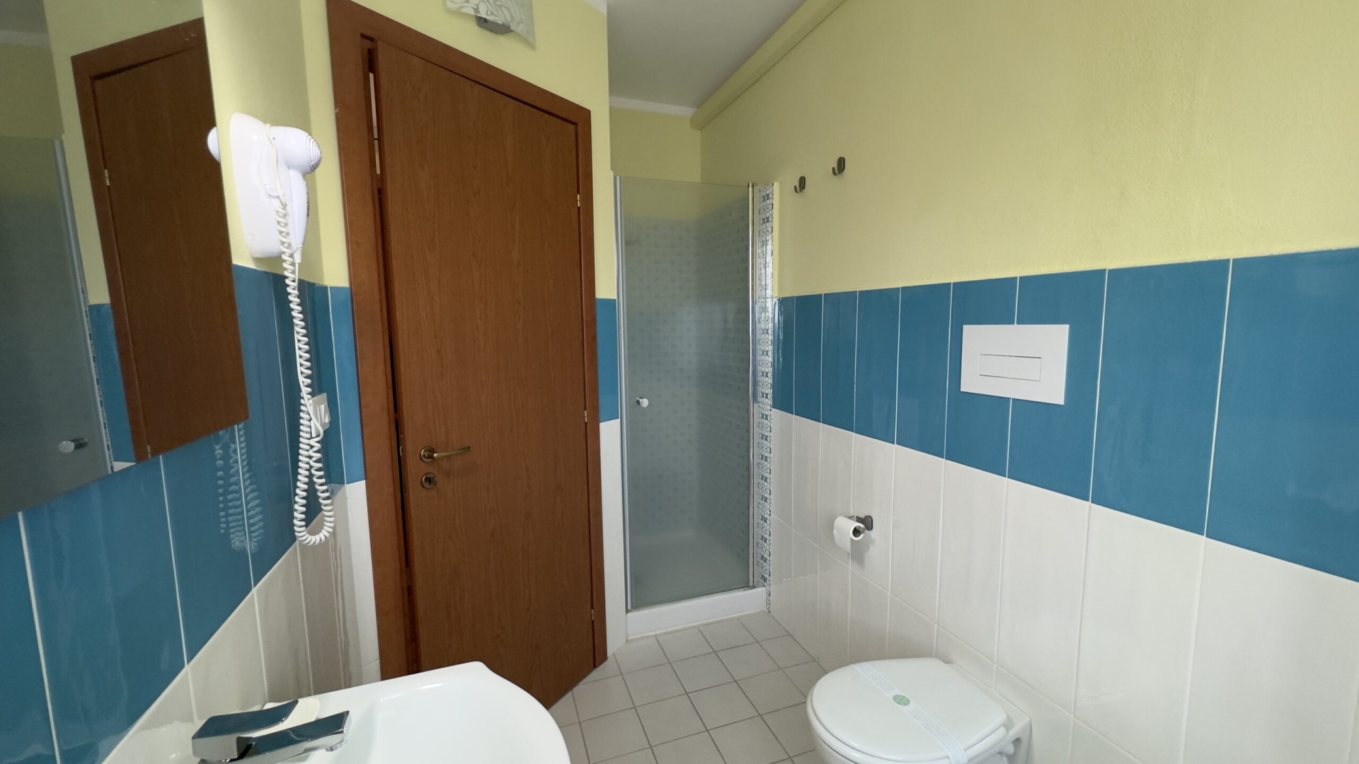 Bagno privato con pareti piastrellate in bianco e azzurro, dotato di un box doccia, WC, e un lavandino con specchio. Asciugacapelli a parete e accessori essenziali per il comfort degli ospiti.