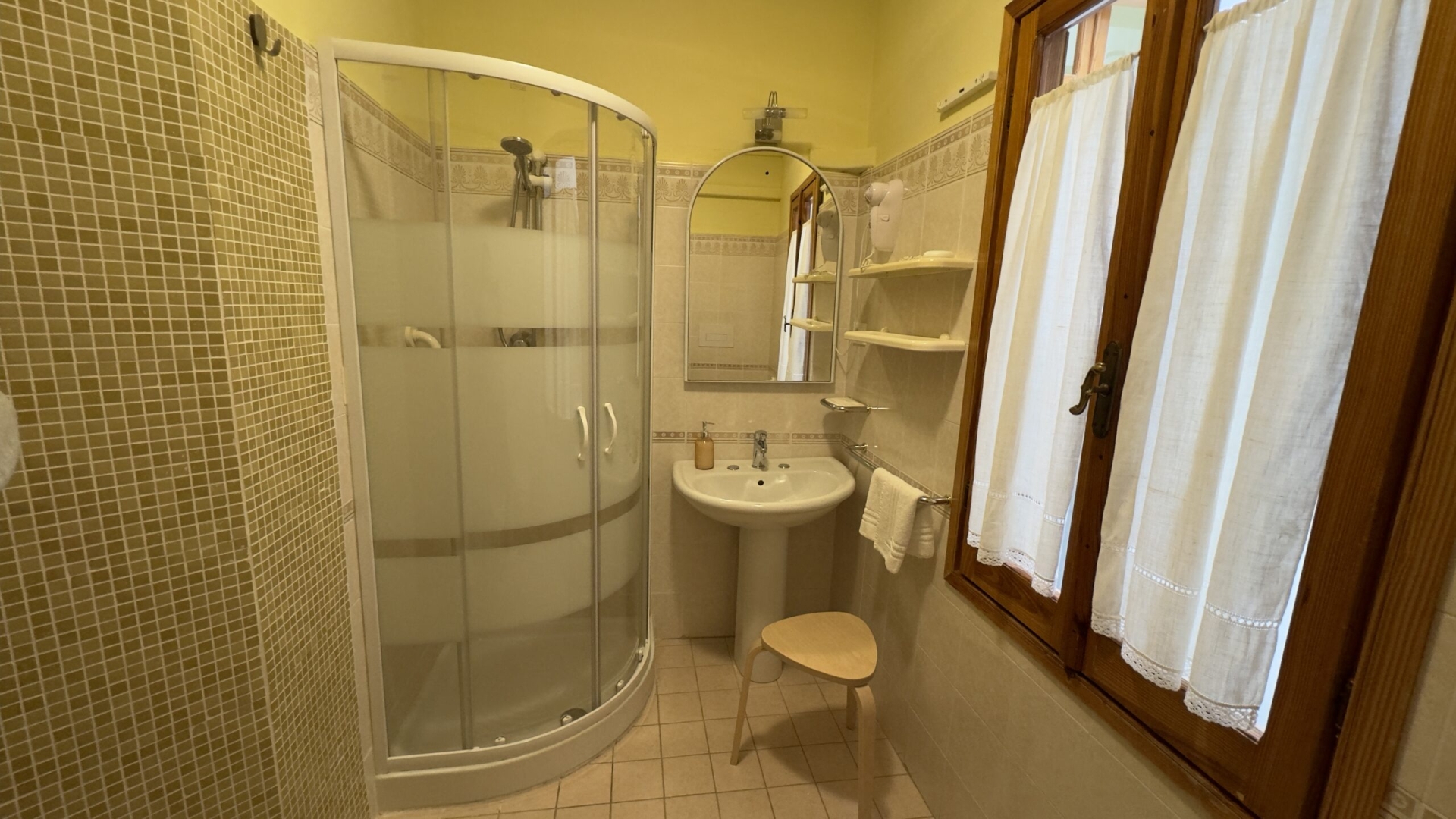 Bagno con box doccia in vetro, lavandino con specchio e ripiani, finestra con tende bianche e asciugacapelli a parete. Piastrelle beige alle pareti e una sedia in legno completano l’arredamento.