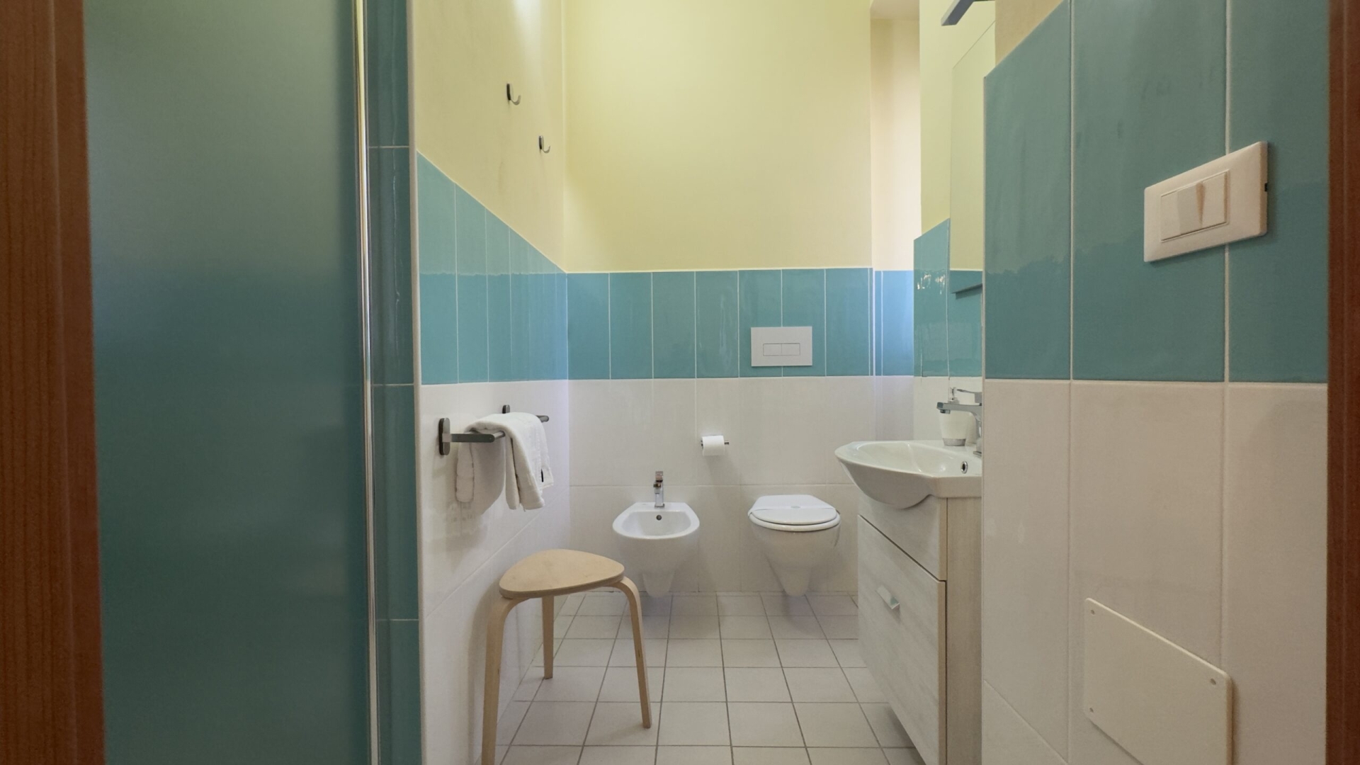 Bagno con piastrelle bianche e azzurre, dotato di bidet, WC, lavandino con mobile bianco e specchio, e un asciugacapelli a parete. Ambiente luminoso e arredamento essenziale.