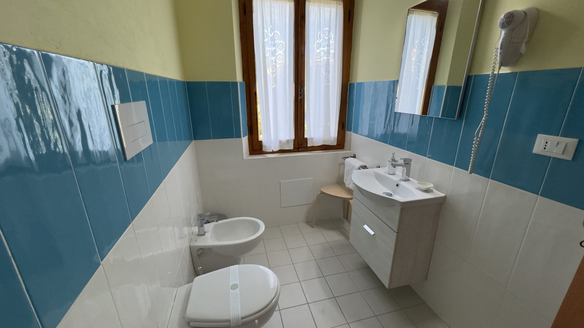 Bagno luminoso con finestra coperta da tende bianche, dotato di WC, bidet, lavandino con specchio e asciugacapelli a parete. Piastrelle azzurre e bianche per un ambiente fresco e accogliente.